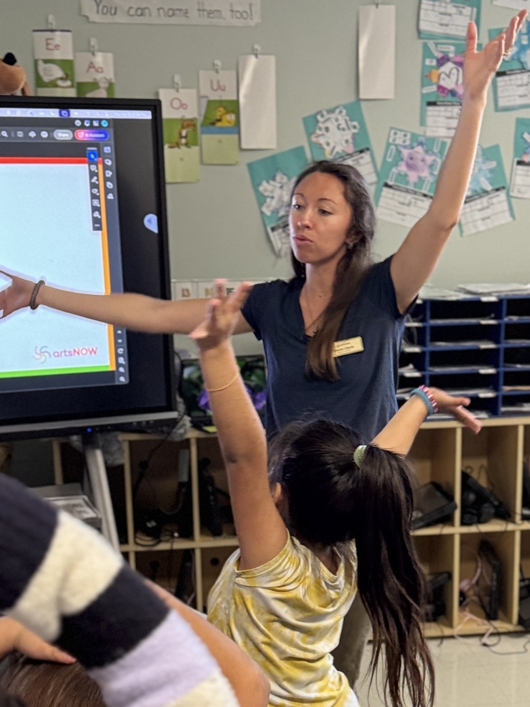 Arts educator leading an interactive lesson with students raising their hands in classroom with digital display
